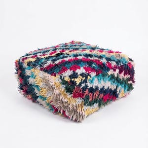 May include: A square floor pouf featuring a thick, shaggy pile texture. The surface displays a vibrant, multi-colored geometric pattern with interwoven threads of deep navy blue, bright magenta, forest green, and pale yellow throughout the design.