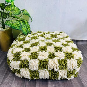 Pode incluir: Um pufe quadrado e tufado com um padrão xadrez de fio verde-oliva e creme. O pufe está colocado sobre um chão de madeira escura, com uma planta em vaso ao fundo.