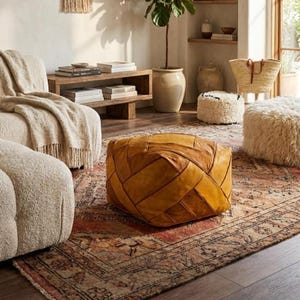 May include: A square, mustard-yellow leather pouf featuring a geometric, woven-style panel construction with visible stitching. The ottoman sits on a patterned area rug in a room with a cream-colored sofa, a wooden shelf, and a potted plant.