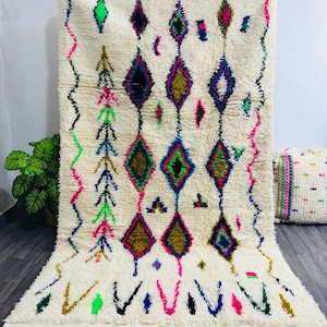 May include: Cream-colored, shaggy rug with a geometric diamond pattern in vibrant colors. The design includes green, pink, blue, and gold accents. The rug has a fringed edge and a matching pillow is visible in the background.