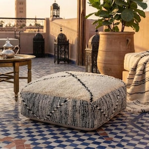 May include: A square floor cushion featuring a thick, cream-colored shaggy top surface and sides patterned with a black and white woven texture. The cushion displays a geometric diamond design created by dark lines across the top and sides.