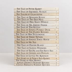 Puede incluir: Una pila vertical de veintitrés libros de tapa dura con lomos de color blanco roto. Cada lomo presenta texto negro con los títulos de Beatrix Potter, incluyendo The Tale of Peter Rabbit, y un número correspondiente del uno al veintitrés a la derecha.