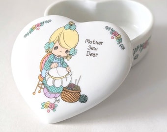 Puede incluir: Caja de porcelana blanca en forma de corazón con un diseño floral. La tapa presenta una ilustración de Precious Moments de una niña cosiendo con el texto "Mother Sew Dear".