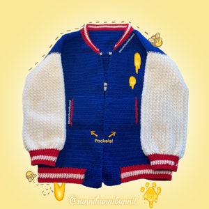 May include: A handmade blue and white crochet varsity jacket with red detailing. The jacket has white sleeves, a blue body, and red trim on the collar, cuffs, and pockets. The jacket has two pockets and the text "Pockets!" is written on the image.