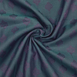 May include: Close-up of teal fabric with a repeating pattern of purple floral shapes. The fabric is draped, showing folds and shadows. The pattern is evenly spaced across the material, creating a textured appearance.