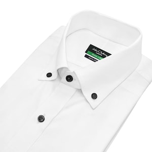 Puede incluir: Una camisa blanca impecable con cuello abotonado y botones oscuros. La camisa está doblada, resaltando su diseño limpio. Una etiqueta con texto es visible en el cuello.