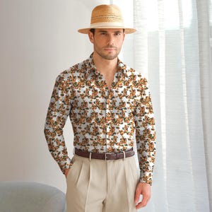 May include: A long-sleeve button-down shirt with a floral pattern in shades of orange and green on a white background. The shirt is paired with khaki pants, a brown belt, and a straw hat. The outfit is casual and stylish.