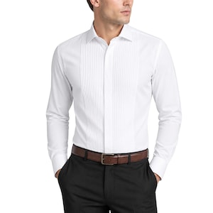 Puede incluir: Una camisa blanca con la parte delantera plisada, mangas largas y cuello clásico. La camisa se combina con pantalones negros y un cinturón de cuero marrón con hebilla plateada. Es una prenda formal.