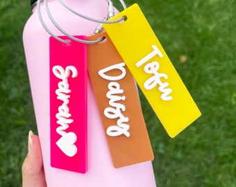 Könnte beinhalten: Eine rosa Wasserflasche mit schwarzem Griff und drei farbigen Namensschildern. Die Anhänger sind pink, braun und gelb, jeweils mit einem Namen in weißer Schrift: Sarah, Daisy und Tofu. Ein weißes Herz befindet sich auf dem Sarah-Anhänger.