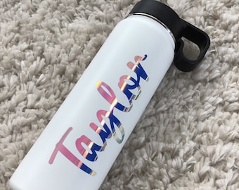 May include: White water bottle with a black lid and handle. The bottle has a holographic sticker with the name "Taylor" in pink and blue.