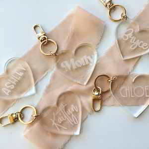 Puede incluir: Cinco llaveros de corazón de acrílico transparente con anillos de llave dorados. Cada llavero tiene un nombre grabado: Ashley, Molly, Kaitlyn, Chloe y Bryce.