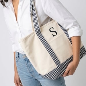 Puede incluir: Bolso tote de lona color crema con ribete y correas de cuadros vichy en blanco y negro. La bolsa presenta la letra negra "S" en el bolsillo delantero. El bolso tote lo sostiene una persona que lleva una camisa blanca y vaqueros azules.
