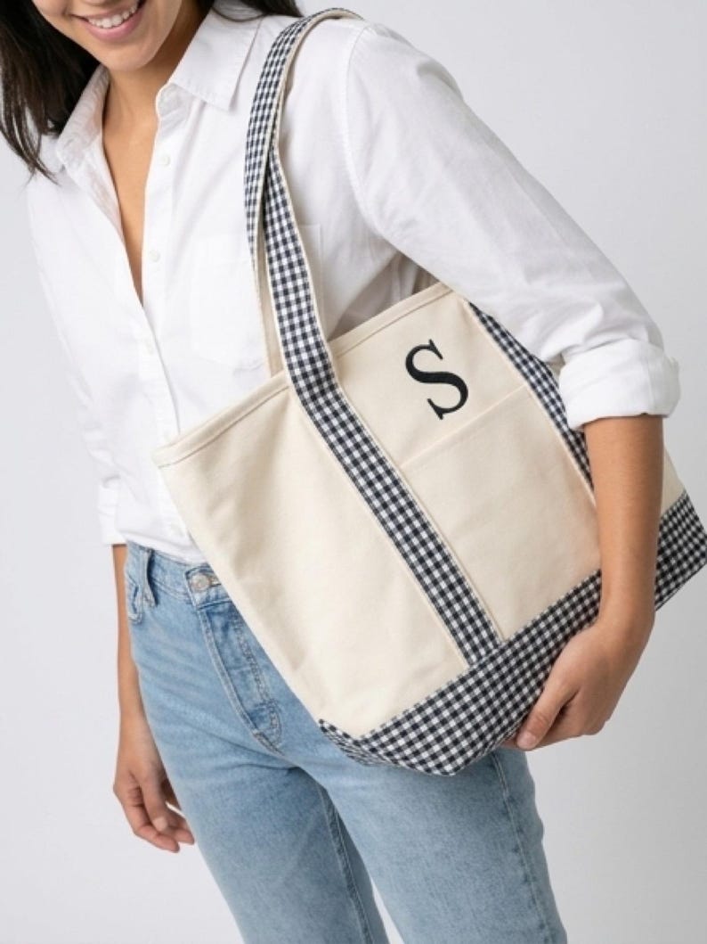 Puede incluir: Bolso tote de lona color crema con ribete y correas de cuadros vichy en blanco y negro. La bolsa presenta la letra negra "S" en el bolsillo delantero. El bolso tote lo sostiene una persona que lleva una camisa blanca y vaqueros azules.