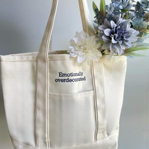 Puede incluir: Bolso tote de lona color crema con el texto bordado "Emotionally overdecorated". El bolso tiene un bolsillo frontal y est&aacute; lleno de flores artificiales blancas y azules. Tiene dos asas largas.