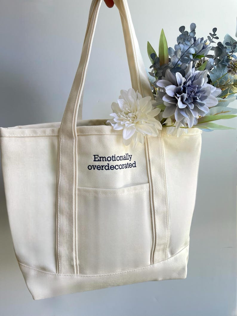 Puede incluir: Bolso tote de lona color crema con el texto bordado "Emotionally overdecorated". El bolso tiene un bolsillo frontal y est&aacute; lleno de flores artificiales blancas y azules. Tiene dos asas largas.