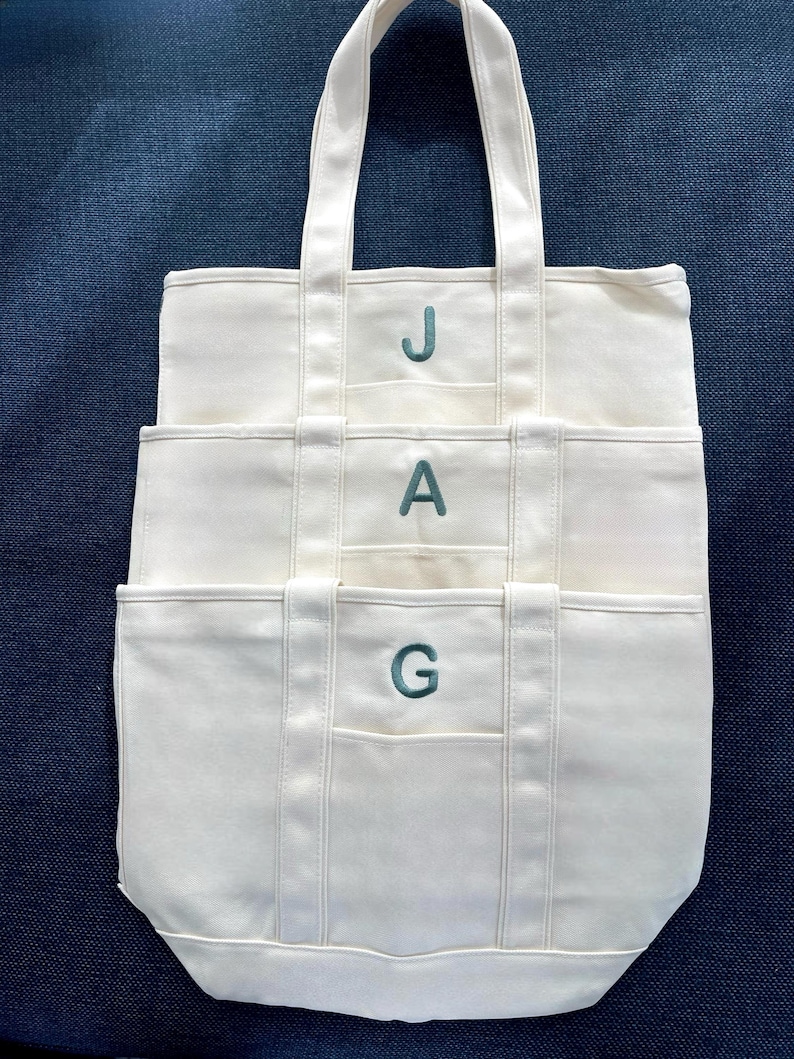 K&ouml;nnte beinhalten: Cremefarbene Canvas-Tragetasche mit drei Taschen und passenden Griffen. Die Buchstaben "JAG" sind in T&uuml;rkis auf die Vorderseite gestickt. Die Tasche liegt auf einer dunkelblauen Oberfl&auml;che.