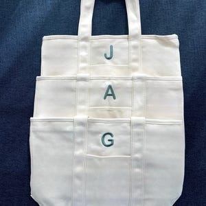 Puede incluir: Bolso tote de lona color crema con tres bolsillos y asas a juego. Las letras "JAG" est&aacute;n bordadas en color turquesa en la parte delantera. El bolso est&aacute; sobre una superficie azul oscuro texturizada.