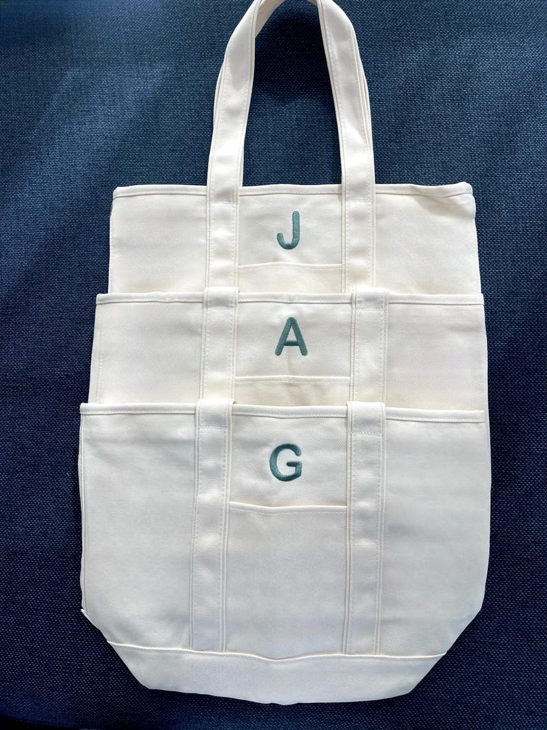 Puede incluir: Bolso tote de lona color crema con tres bolsillos y asas a juego. Las letras "JAG" est&aacute;n bordadas en color turquesa en la parte delantera. El bolso est&aacute; sobre una superficie azul oscuro texturizada.