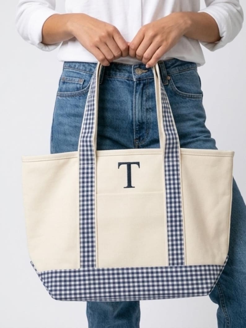 Puede incluir: Bolso tote de lona color crema con ribete y asas de cuadros vichy azul marino. La bolsa presenta la letra "T" azul marino en el bolsillo delantero. El bolso tote lo sostiene una persona que lleva vaqueros azules y una camisa blanca.