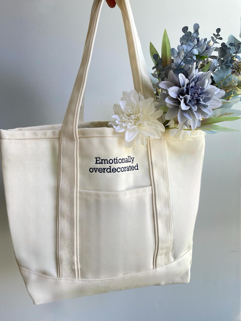 K&ouml;nnte beinhalten: Cremefarbene Canvas-Tragetasche mit dem gestickten Text "Emotionally overdecorated". Die Tasche hat eine Vordertasche und ist mit wei&szlig;en und blauen Kunstblumen gef&uuml;llt. Sie hat zwei lange Griffe.