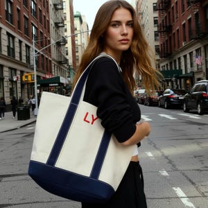 K&ouml;nnte beinhalten: Eine cremefarbene Canvas-Tragetasche mit marineblauen Bes&auml;tzen und Tr&auml;gern, auf der die roten Buchstaben "LY" zu sehen sind. Die Tasche wird von einer Person in einer Stadtstra&szlig;e getragen, mit Geb&auml;uden im Hintergrund.