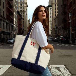Puede incluir: Un bolso tote de lona grande con cuerpo color crema, ribetes y correas azul marino. La bolsa presenta las letras rojas "LY" en la parte delantera. La bolsa se lleva en una calle de la ciudad.