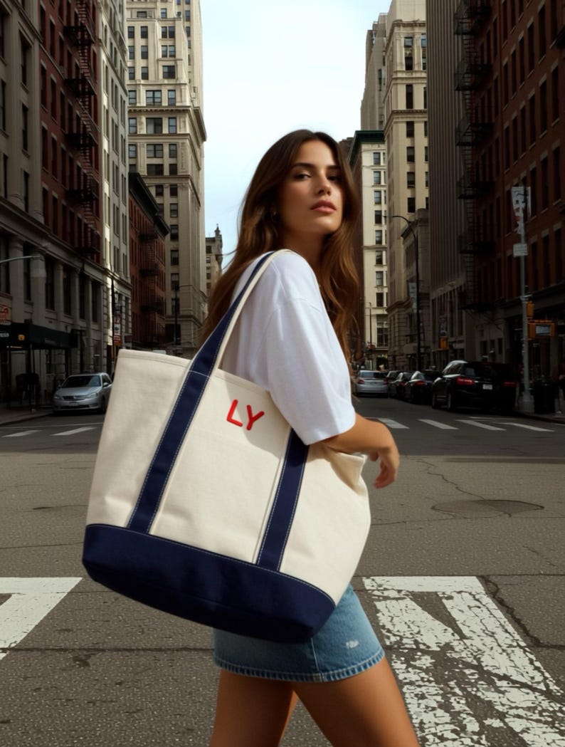 Puede incluir: Un bolso tote de lona grande con cuerpo color crema, ribetes y correas azul marino. La bolsa presenta las letras rojas "LY" en la parte delantera. La bolsa se lleva en una calle de la ciudad.