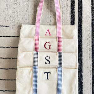 Puede incluir: Bolso tote de lona color crema con m&uacute;ltiples bolsillos y un estampado de cuadros vichy. El bolso presenta detalles de cuadros vichy rosas, rojos, negros y azules en las asas y el ribete. Bordado con las letras A, G, S y T.