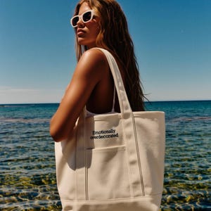 K&ouml;nnte beinhalten: Cremefarbene Canvas-Tragetasche mit dem Text "Emotionally overdecorated" in Schwarz. Die Tasche hat zwei Au&szlig;entaschen und eine rechteckige Form. Der Hintergrund zeigt eine Person mit Sonnenbrille in der N&auml;he des Meeres.