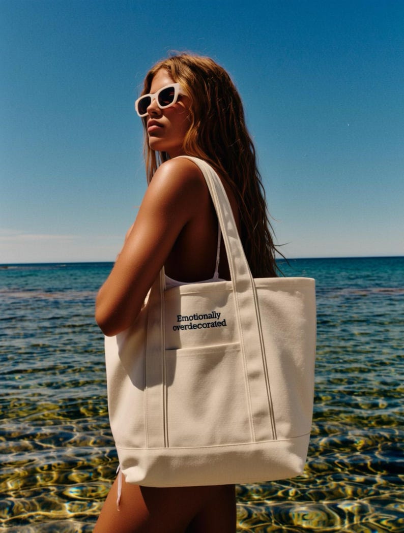 K&ouml;nnte beinhalten: Cremefarbene Canvas-Tragetasche mit dem Text "Emotionally overdecorated" in Schwarz. Die Tasche hat zwei Au&szlig;entaschen und eine rechteckige Form. Der Hintergrund zeigt eine Person mit Sonnenbrille in der N&auml;he des Meeres.