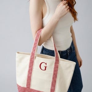 Puede incluir: Bolso tote de lona color crema con ribete y asas de cuadros rojos. Presenta la letra "G" bordada en rojo en el bolsillo delantero. Un accesorio elegante y pr&aacute;ctico.