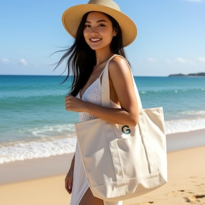 Puede incluir: Un bolso tote color crema con una 'G' verde bordada en el bolsillo delantero. El bolso tiene forma rectangular con una correa para el hombro. La imagen muestra el bolso en una playa con una mujer con sombrero y vestido blanco.