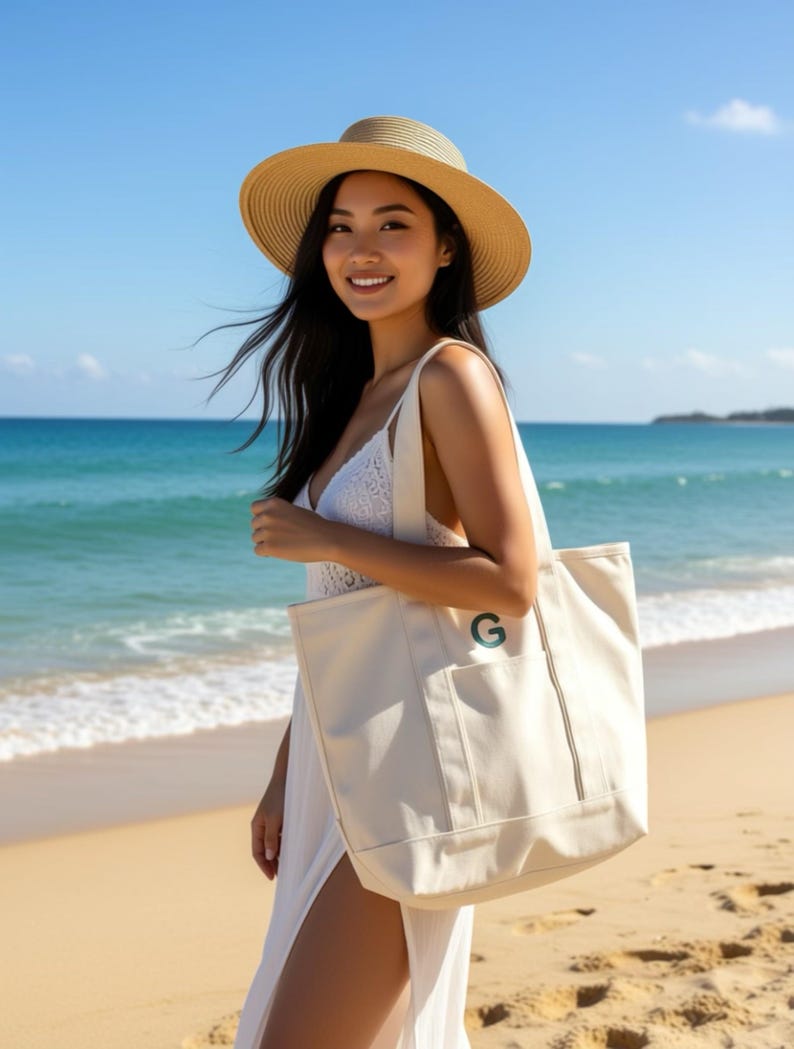 Puede incluir: Un bolso tote color crema con una 'G' verde bordada en el bolsillo delantero. El bolso tiene forma rectangular con una correa para el hombro. La imagen muestra el bolso en una playa con una mujer con sombrero y vestido blanco.