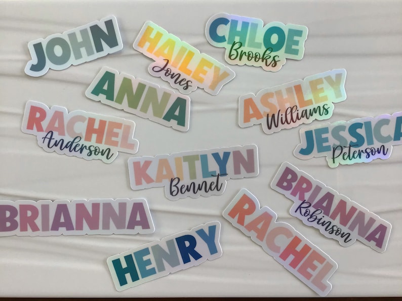 May include: A collection of 12 iridescent rainbow name stickers with various names including John, Hailey, Chloe, Anna, Rachel, Ashley, Jessica, Kaitlyn, Brianna, Henry, and Rachel. Each sticker has a different color gradient and a last name.