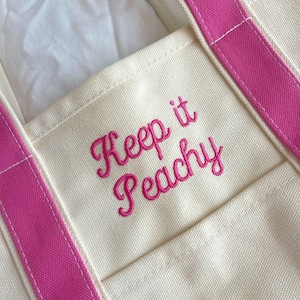 Puede incluir: Bolso tote color crema con correas rosa brillante y la frase bordada "Keep it Peachy" en cursiva. El bolso tiene un bolsillo en la parte delantera y est&aacute; hecho de un material de lona resistente.