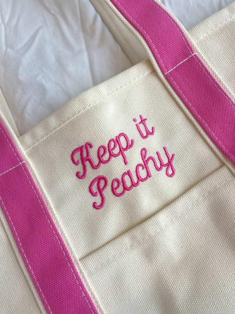 Puede incluir: Bolso tote color crema con correas rosa brillante y la frase bordada "Keep it Peachy" en cursiva. El bolso tiene un bolsillo en la parte delantera y est&aacute; hecho de un material de lona resistente.
