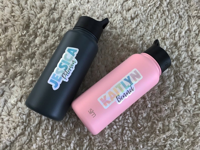 May include: Two water bottles with personalized stickers. The black bottle has a sticker with the name "Jessica Peterson" in a rainbow gradient font. The pink bottle has a sticker with the name "Kaitlyn Bennet" in a rainbow gradient font.