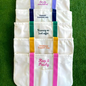 K&ouml;nnte beinhalten: Eine Ansammlung von cremefarbenen Canvas-Tragetaschen mit farbigen Akzenten. Jede Tasche hat einen anderen gestickten Spruch: "Tote-ally overwhelmed", "Dramatic Essentials only", "Running on Iced coffee", "Emotional Support Tote" und "Keep it Peachy".