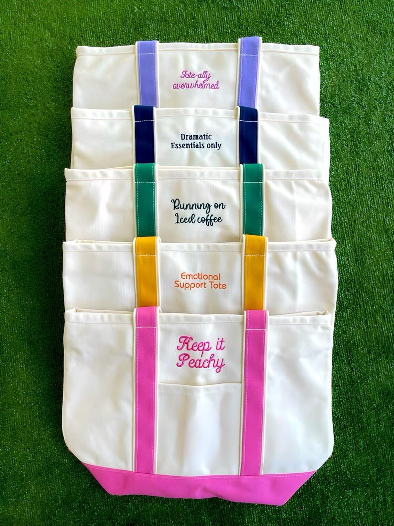 K&ouml;nnte beinhalten: Eine Ansammlung von cremefarbenen Canvas-Tragetaschen mit farbigen Akzenten. Jede Tasche hat einen anderen gestickten Spruch: "Tote-ally overwhelmed", "Dramatic Essentials only", "Running on Iced coffee", "Emotional Support Tote" und "Keep it Peachy".