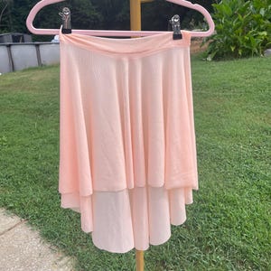 Op de afbeelding: Lichtperzikroze balletrok met een gelaagd ontwerp, hangend aan een roze hanger. De rok heeft een vloeiend, gelaagd silhouet, perfect voor dans of kostuums. Gemaakt van een zachte, lichte stof.