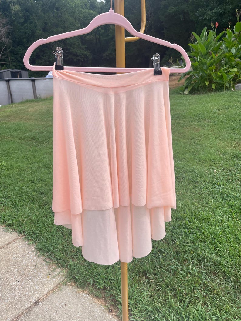 Op de afbeelding: Lichtperzikroze balletrok met een gelaagd ontwerp, hangend aan een roze hanger. De rok heeft een vloeiend, gelaagd silhouet, perfect voor dans of kostuums. Gemaakt van een zachte, lichte stof.