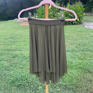 Op de afbeelding: Een olijfgroene rok met een high-low zoom hangt aan een roze hanger. De rok is gemaakt van een lichte, vloeiende stof. De achtergrond toont groen gras en gebladerte.