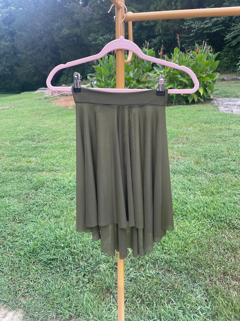 Op de afbeelding: Een olijfgroene rok met een high-low zoom hangt aan een roze hanger. De rok is gemaakt van een lichte, vloeiende stof. De achtergrond toont groen gras en gebladerte.