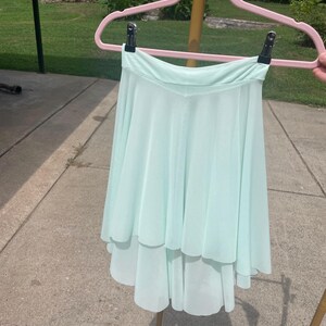 Op de afbeelding: Een lichtgroene, gelaagde dansrok hangend aan een roze hanger. De rok heeft een vloeiend, asymmetrisch ontwerp, met een kortere voorkant en een langere achterkant. De stof lijkt licht en luchtig, geschikt voor dans.