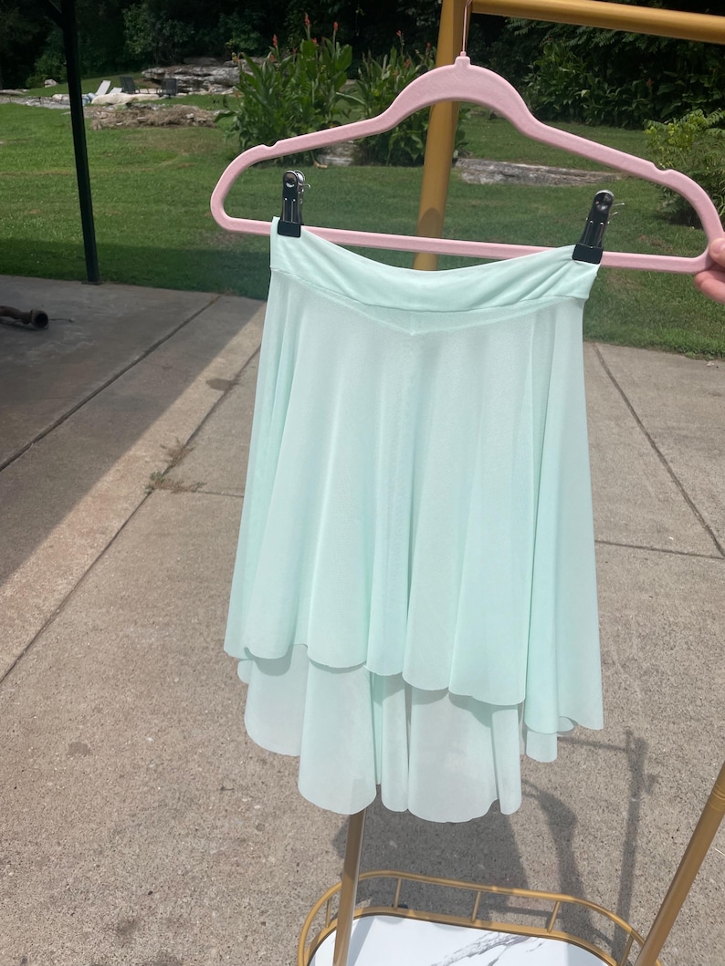 Op de afbeelding: Een lichtgroene, gelaagde dansrok hangend aan een roze hanger. De rok heeft een vloeiend, asymmetrisch ontwerp, met een kortere voorkant en een langere achterkant. De stof lijkt licht en luchtig, geschikt voor dans.