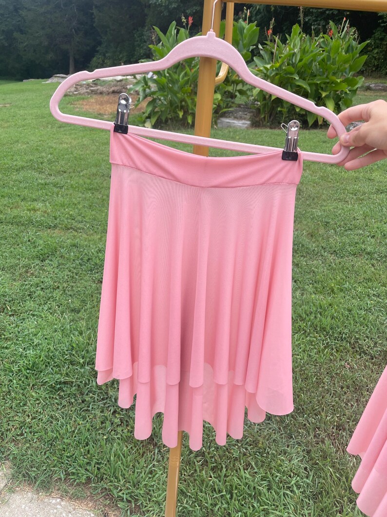 Op de afbeelding: Een lichtroze, gelaagde dansrok wordt getoond op een roze hanger. De rok heeft een getailleerde tailleband en een vloeiende, asymmetrische zoom. De rok is gemaakt van een lichtgewicht, transparante stof en hangt buiten op een zonnige dag.