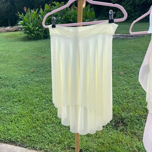 Op de afbeelding: Een lichtgele dansrok met een gelaagde, asymmetrische zoom hangt aan een roze hanger. De rok is gemaakt van een lichtgewicht, vloeiende stof. De achtergrond toont groen gras en gebladerte.