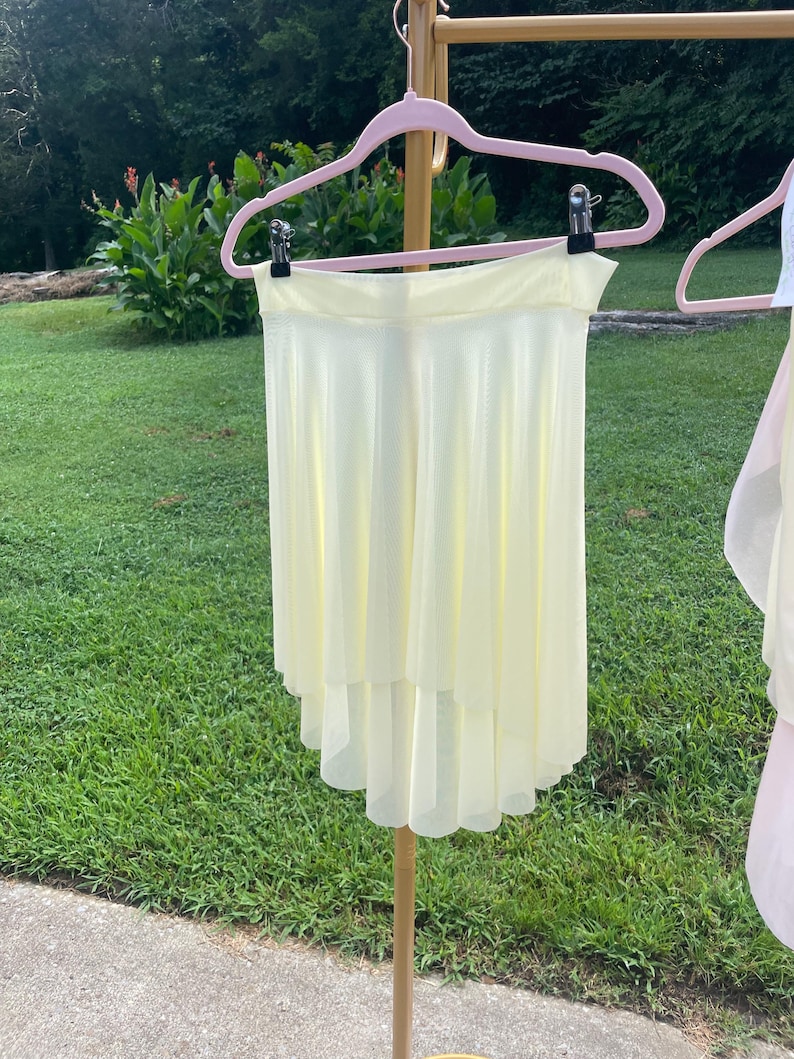 Op de afbeelding: Een lichtgele dansrok met een gelaagde, asymmetrische zoom hangt aan een roze hanger. De rok is gemaakt van een lichtgewicht, vloeiende stof. De achtergrond toont groen gras en gebladerte.
