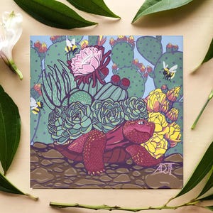 May include: A square art print featuring a maroon tortoise with a shell composed of green succulents, a large pink cactus flower, and yellow blossoms. The scene includes a prickly pear cactus background, flying bees, and a rocky ground with the signature ADJ 2022.