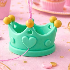 May include: A festive party scene with pink and gold accents. A teal crown-shaped centerpiece is surrounded by cupcakes, plates, and a pink tablecloth. Purple and pink balloons float above the table, creating a celebratory atmosphere.